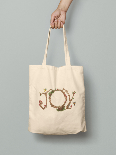 Hand holding a cream colored tote bag with the illustrated word JOY